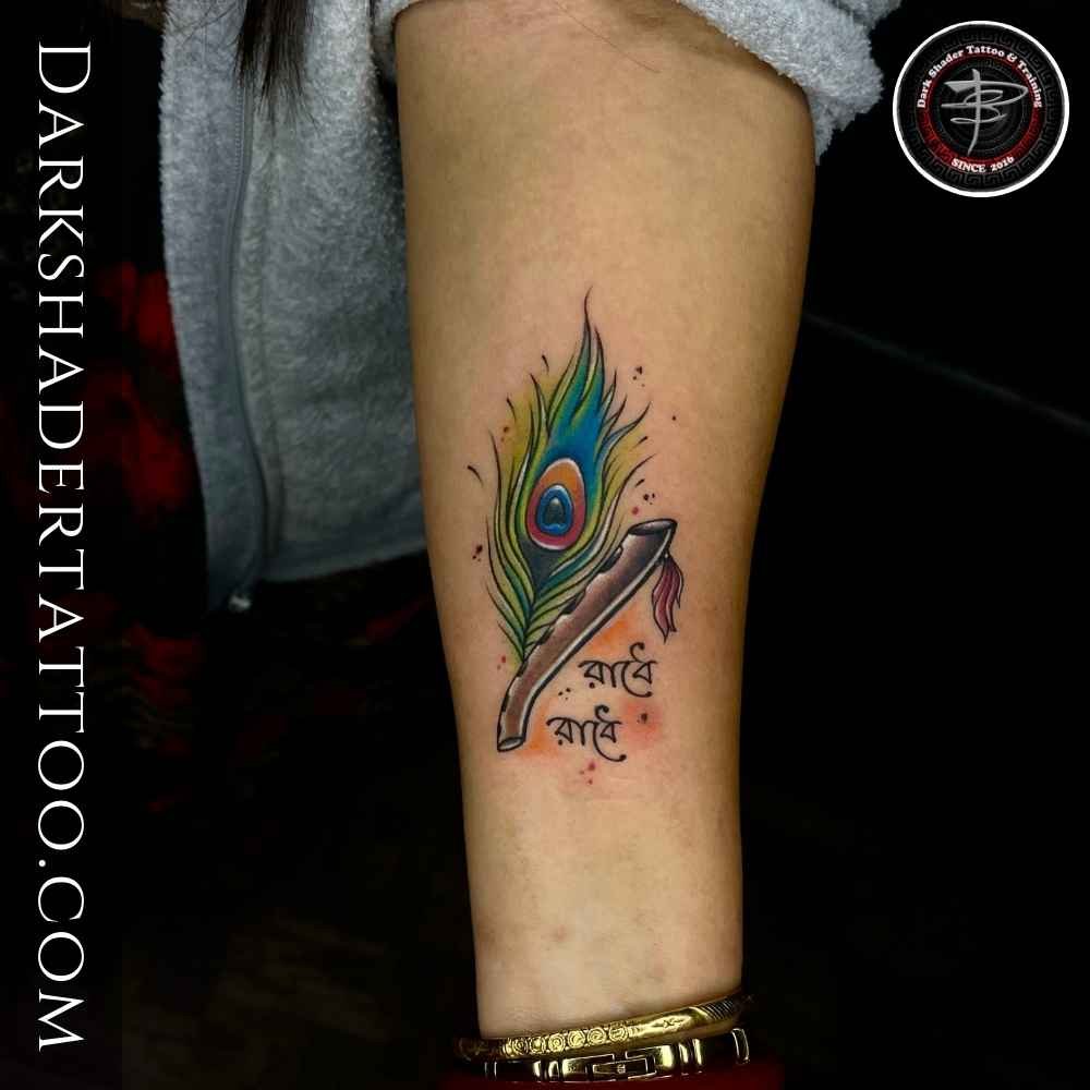 A "Radhe Radhe" tattoo in Bengali with a peacock feather and Krishna’s flute on a girl’s wrist.