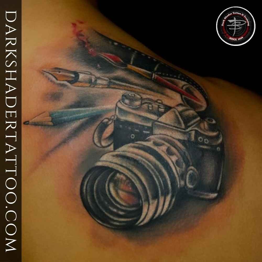 A tattoo of a camera with pencil, pen, reel, and brush combined, designed on a girl’s upper back below the neck.