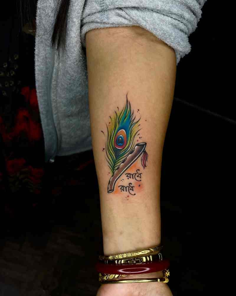 Krishna feather tattoo with "Radhe Radhe" written in Bengali, beautifully designed with green shades on a girl's hand.