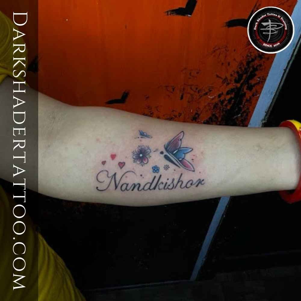 "Nandakishor" text tattoo on a girl’s wrist with beautiful additions like a butterfly, heart, and flower.