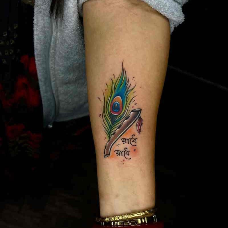 Krishna feather tattoo with "Radhe Radhe" written in Bengali, beautifully designed with green shades on a girl's hand.