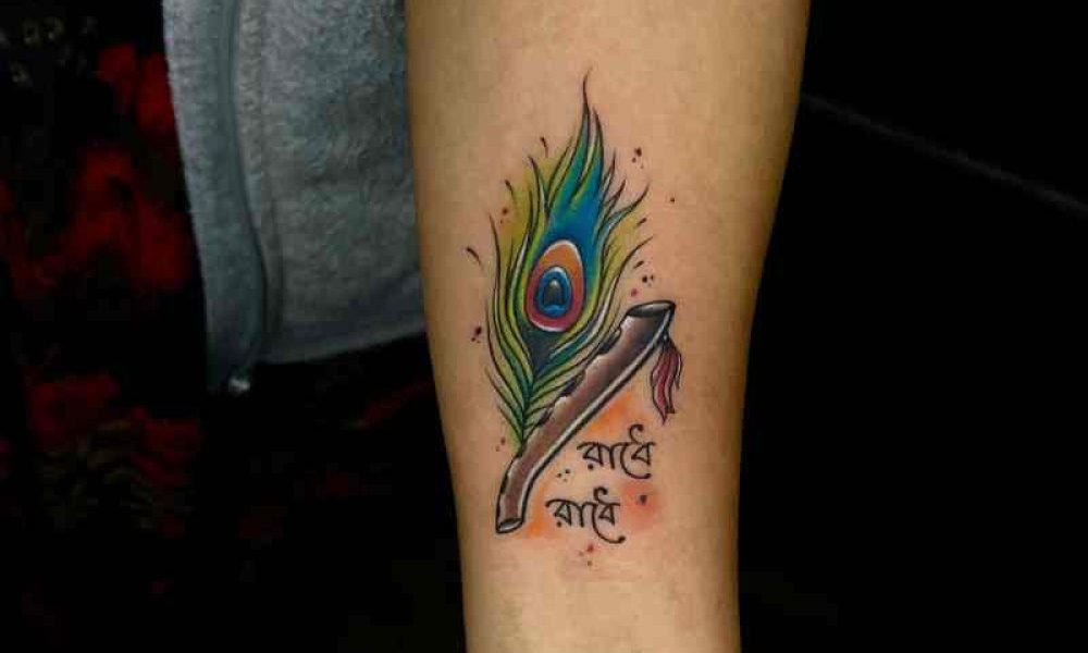 Krishna feather tattoo with "Radhe Radhe" written in Bengali, beautifully designed with green shades on a girl's hand.