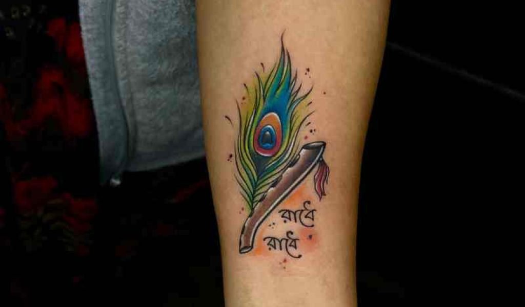 Krishna feather tattoo with "Radhe Radhe" written in Bengali, beautifully designed with green shades on a girl's hand.